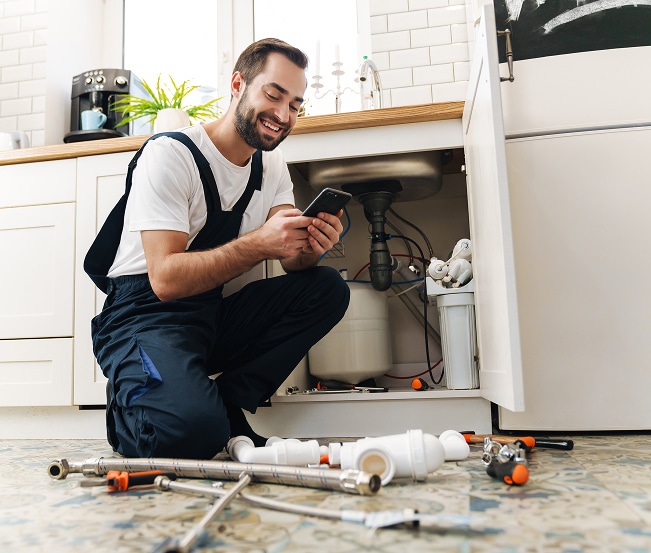 Plombier souriant en salopette accroupi, consulte son smartphone devant un évier de cuisine. Des outils de plomberie sont au sol.
