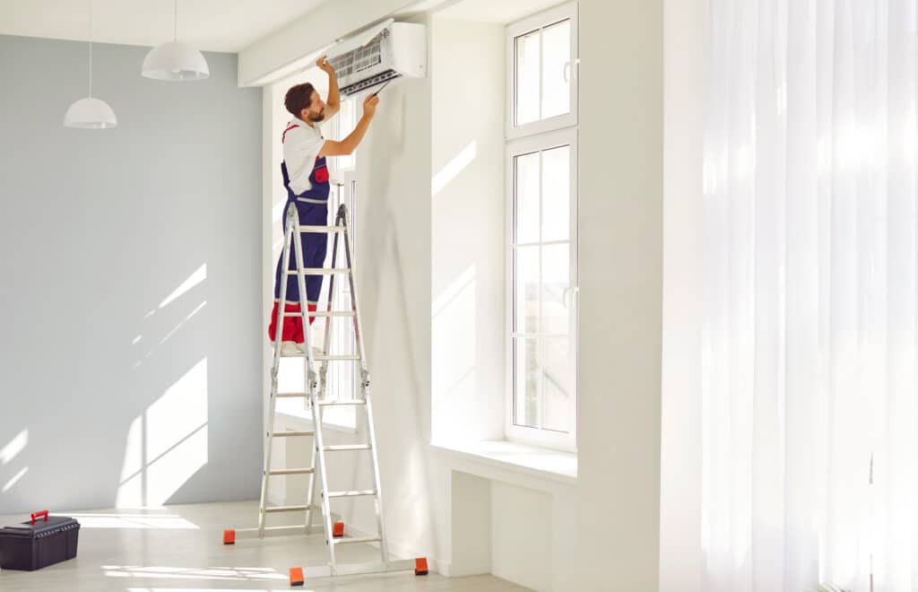 Technicien souriant sur une échelle entretient un climatiseur mural blanc. Pièce lumineuse avec fenêtres, rideaux et ombres solaires au sol.
