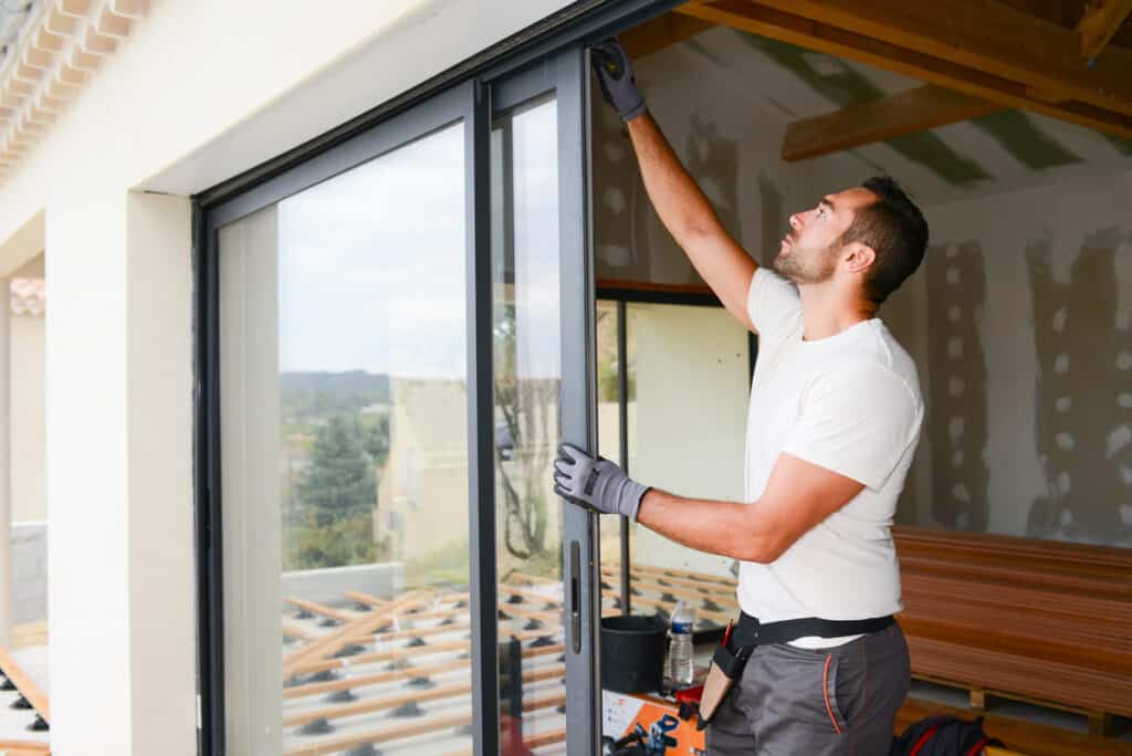 Homme en gants installant ou ajustant une grande porte-fenêtre coulissante en verre sur une terrasse de maison en construction.