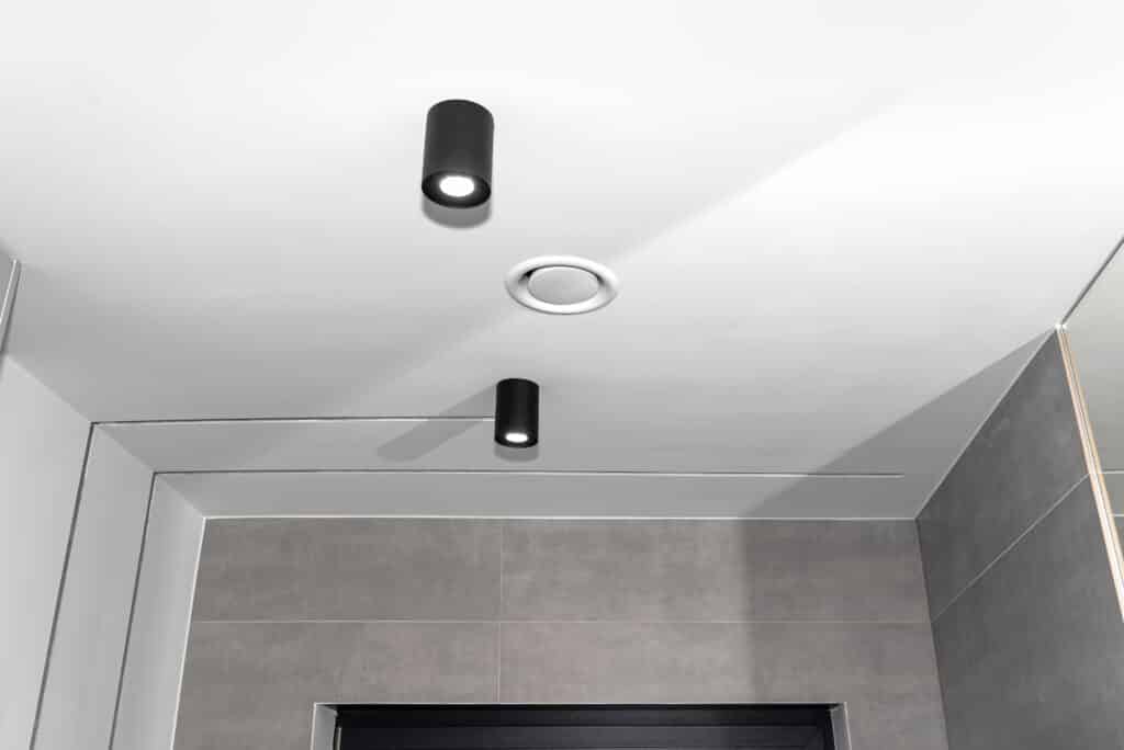 Plafond blanc moderne avec deux spots noirs cylindriques allumés et une aération ronde. Murs carrelés gris, ambiance minimaliste.