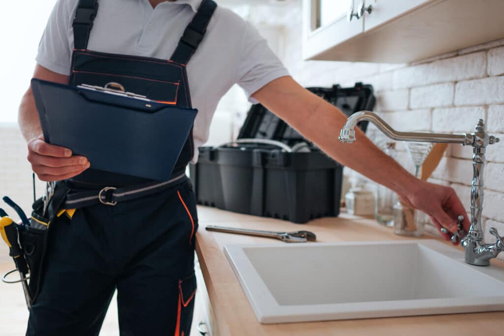 Réparation robinet cuisine: Plombier expert à domicile Plombier en salopette tenant un clipboard, ajustant un robinet d'évier de cuisine en chrome. Une boîte à outils est visible.