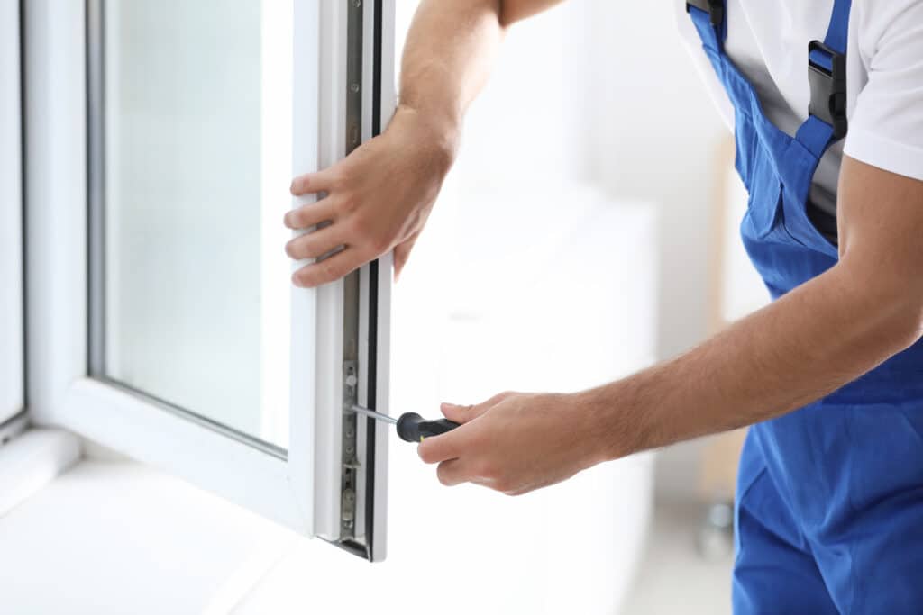 Réglage & Réparation Fenêtre PVC : Service Professionnel Technicien en salopette bleue réglant le mécanisme d'une fenêtre PVC blanche avec un tournevis, mains en action.